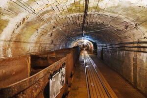 The image captures a vintage mining cart resting on rusty tracks in a dark mine tunnel. The atmosphere is mysterious, with a brick ceiling and shadows creating an eerie ambiance, gold mine photo