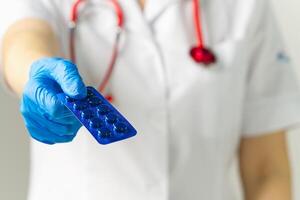 A doctor in a white coat and stethoscope inspects a blister pack of blue, round pills in a clinical setting, showcasing professionalism and careful attention to medication management. photo