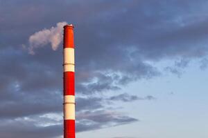 A tall industrial chimney with a bold red-and-white striped pattern emits thick white smoke into a cloudy sky. Sunlight breaks through the clouds, creating a dramatic ambiance around the scene. photo