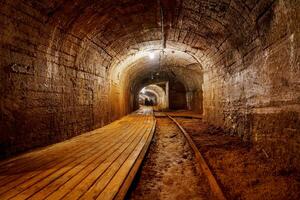 A dimly lit tunnel features a worn wooden walkway and rusty railroad tracks. The rough stone walls add to the dark, mysterious atmosphere of this abandoned passage, cooper mine photo