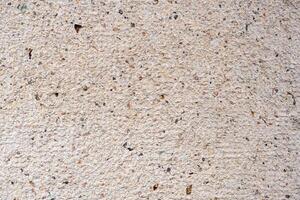 Close-up of a textured surface, likely concrete or a wall, with specks of black, brown, and red. The overall color is light beige or off-white. photo