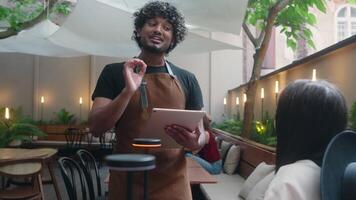 Friendly restaurant worker guy Arabian Indian male waiter in apron serve woman client in cafe customer service smiling man serving in cafeteria communicate with visitor write food order digital tablet video