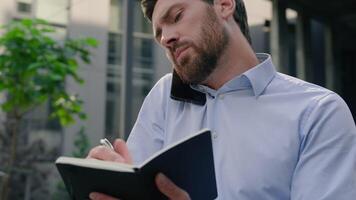 Serious Caucasian male talking mobile phone discuss project consulting client talk smartphone man businessman guy business chat solve problem writing notes in notebook write idea information in city video