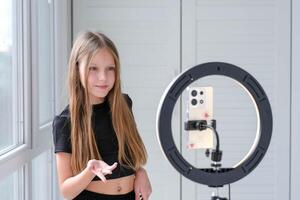 Beautiful teenage girl posing in front of a phone on a tripod. Child blogger shoots in front of camera and ring lamp photo