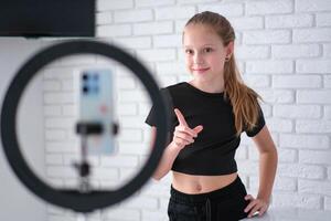 Beautiful teenage girl posing in front of a phone on a tripod. Child blogger shoots in front of camera and ring lamp photo