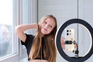 Beautiful teenage girl posing in front of a phone on a tripod. Child blogger shoots in front of camera and ring lamp photo