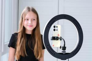 Beautiful teenage girl posing in front of a phone on a tripod. Child blogger shoots in front of camera and ring lamp photo