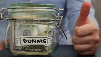 Female hands showing thumb up like sign Saving Money In Glass Jar filled with Dollars banknotes. DONATE transcription in front of jar. Managing personal finances extra income for future insecurity background video