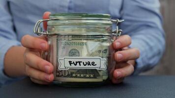 Unrecognizable woman put on table Saving Money In Glass Jar filled with Dollars banknotes. FUTURE transcription in front of jar. Managing personal finances extra income for future insecurity background video