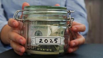 Unrecognizable woman put on table Saving Money In Glass Jar filled with Dollars banknotes. 2025 year transcription in front of jar. Managing personal finances extra income for future insecurity background video