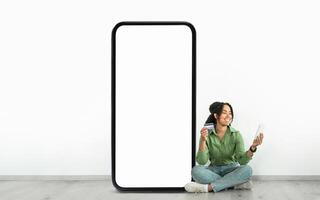 A woman is sitting on the floor, smiling as she holds a credit card and a tablet, next to a large blank smartphone display. The setting suggests a relaxed digital shopping experience. photo