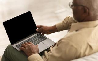Back View Of Senior Black Man Using Laptop With Empty Computer Screen For Website Advertisement Sitting Working Online Indoors. High Angle View. Mockup, Selective Focus On Display photo