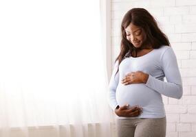 Peaceful pregnant black woman hugging her tummy next to window at home, free space photo