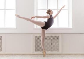 Arabesque. Classical Ballet dancer side view. Beautiful graceful ballerine in black practice ballet position near large window in light hall. Ballet class training, high-key soft toning. photo