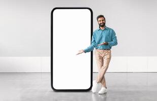 Middle eastern man in a blue shirt and beige pants stands next to a large blank smartphone screen, gesturing as if inviting viewers to engage with the display in a contemporary setting. photo