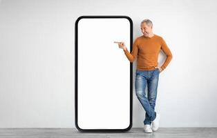 A man in a brown sweater and jeans stands next to a large, empty smartphone display, posing with a playful gesture. The setting is bright and minimalistic, emphasizing the display. photo