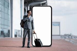 African American man in a suit stands at the airport terminal next to huge phone with blank screen, talking on his smartphone while holding a suitcase. photo