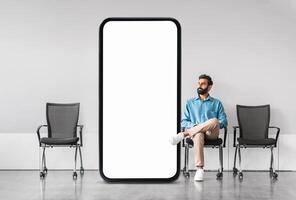 Arab man casually sits next to a large blank screen in a contemporary waiting area, creating an intriguing contrast in space and emphasizing the minimalist design of the environment. photo
