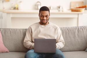 Portrait of bearded young black male freelancer working from home, sitting on couch, using laptop, copy space. Relaxed African American guy enjoying weekend, websurfing on notebook photo