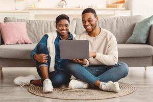 People And Technology. Portrait of positive black family of two people using laptop computer, watching or movie, shopping in online store, sitting on the floor carpet in living room at home photo