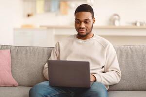 Modern Remote Job Lifestyle. Portrait of black young man using laptop sitting on the sofa at home, working online, browsing internet or watching on personal computer, free copy space photo
