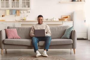 Modern Remote Job Lifestyle. Smiling black young man using laptop sitting on sofa at home, working online, browsing internet or watching on computer, free space. People And Technology photo