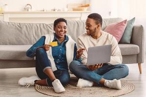 Online Shopping From Home, E-commerce Concept. Happy young African American couple sitting on floor carpet at home and buying clothes or products online, paying with credit card using laptop photo