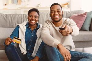 On Demand Concept. Smiling African American millennial couple sitting on floor, guy using tv remote controller, lady holding golden credit card, paying for vod service, multimedia streaming photo