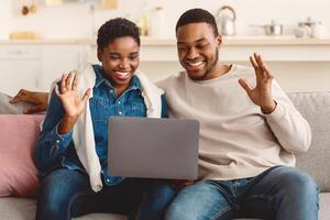 Online Communication. Smiling african american man and woman making digital chat with friends or family using laptop, greeting and waving to webcam, sitting on the couch in living room photo