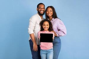 Portrait of happy family of three African American people showing digital tablet with black empty screen, smiling girl holding device with blank space for mockup, standing over blue studio background photo