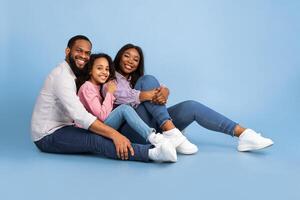 Cool Team. Beautiful and happy smiling young black family hugging and having fun time together while sitting on the floor and looking at camera, isolated on blue studio background, free copy space photo