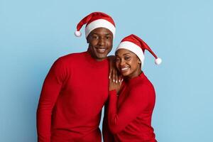 A joyful black couple poses together wearing matching red sweaters and festive Santa hats. They smile warmly against a light blue background, radiating holiday cheer. photo