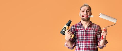 Multitasking problem. Desperate repairman holding perforator, paint roller and brush simultaneously, yellow studio background photo
