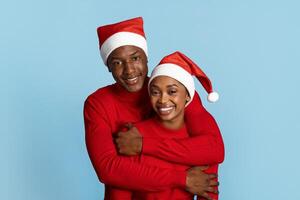 A joyful black couple embraces against a blue background, both wearing matching red sweaters and festive Santa hats. They radiate happiness and togetherness during the holiday season. photo