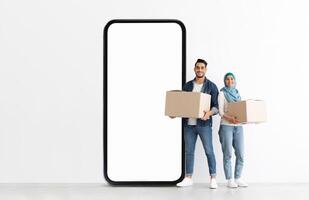 A young Muslim man and woman stand together, smiling while holding cardboard boxes. They are positioned next to a large empty smartphone display in a bright, minimalist space. photo