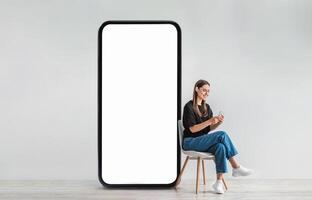 A young woman sits comfortably on a chair, engaging with her smartphone while a large blank screen stands next to her in a contemporary space, suggesting a digital connection. photo