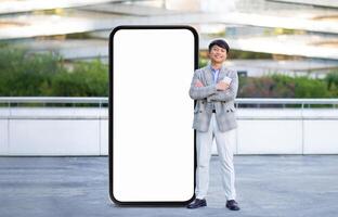 A young Asian man professional smiles and crosses his arms while standing next to a large blank digital display in a sleek urban environment photo