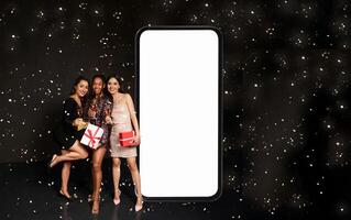 Three smiling friends pose together at a festive event, each holding colorful gifts in a setting adorned with sparkling effects and a blank phone screen behind them. photo
