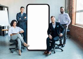 A group of four professionals stands confidently in a contemporary office, showcasing a large blank smartphone screen. Their attire reflects a blend of formality and style, emphasizing teamwork. photo