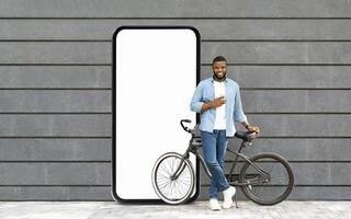 A young black man stands casually with a smartphone in hand, leaning against his bike, next to a large blank screen resembling a smartphone, set against a modern wall. photo