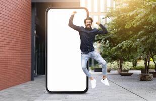 Indian man dressed in casual attire jumps joyfully in front of a large blank smartphone screen, situated in a sunny outdoor setting with greenery and modern architecture. photo