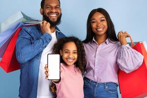 Online Shopping App. Closeup portrait of happy black family holding shopping bags, smiling girl showing cellphone with white empty isolated screen in hand. Blank space for mock up, blue studio wall photo