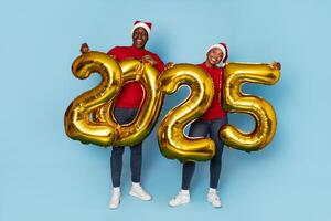 Two black friends celebrate the arrival of 2025, dressed in festive red outfits and Santa hats. They proudly hold large golden numbers, creating a joyful and cheerful atmosphere. photo