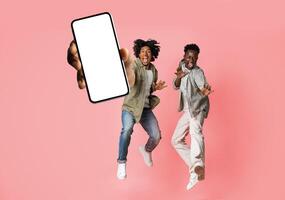 Excited black male friends jumping and demonstrating smartphone with white blank screen, african american guys offering app or website, pink studio background, mockup collage banner photo