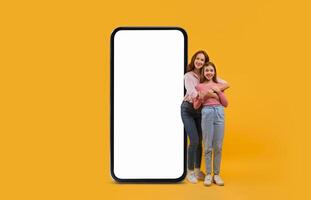 A mother and daughter are happily standing together, embracing each other, beside a large blank smartphone. The bright yellow background enhances their joyful expression. photo