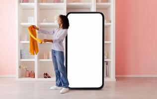 A woman is holding an orange shirt while standing beside a large smartphone display in a trendy interior. The setting features shelves with stylish items and a pink backdrop. photo
