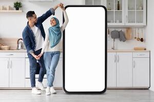 A joyful Muslim couple dances in their contemporary kitchen, playfully interacting around a large smartphone frame while enjoying their time together in a bright, inviting space. photo