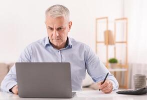 Budget Planning. Pensive Senior Man Using Laptop And Writing In Notebook Sitting At Desk At Home, Empty Space photo