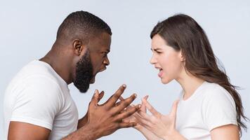 Multiracial couple quarreling, shouting at each other, having relationship problems, standing together over light background, side view photo