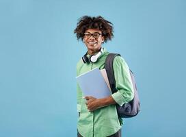 Smart african american student guy with backpack and notebooks smiling at camera, posing over blue studio background. Education, university, college, studying, course concept photo